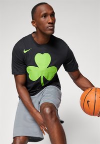 Camiseta negra de Nike con un gran logo de trébol verde, combinada con pantalones cortos grises. El modelo sostiene un balón de baloncesto.