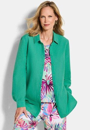 Frau mit blonden Haaren, die ein hellgrünes Hemd mit Knöpfen über einem bunt gemusterten, floralen Oberteil und passenden, floralen Hosen trägt, posiert mit einer Hand in der Tasche.