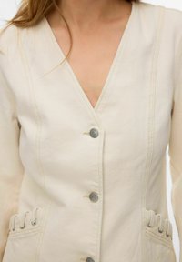 Femme portant une veste beige boutonnée avec col en V et un détail de tissu froncé près de la taille des deux côtés.