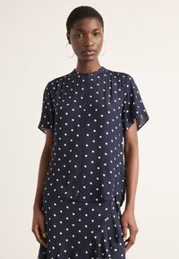 Tumed blouse valgete täppidega, lühikesed varrukad, nööpidega esikülg, pehme kangas ja lõdvestatud lõige, kombineeritud sobiva täppidega seelikuga.