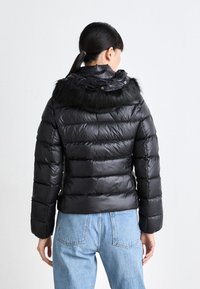Giubbotto piumino nero con cappuccio foderato in pelliccia, design trapuntato e texture lucida, abbinato a jeans azzurri chiari.