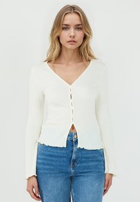 Top bianco a maniche lunghe con scollo a V, con chiusura a bottoni davanti e orlo leggermente sfrangiato, abbinato a jeans in denim blu.