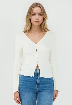 Haut à manches longues blanc côtelé avec décolleté en V, présentant une fermeture à boutons sur le devant et un ourlet légèrement effiloché, associé à un jean en denim bleu.