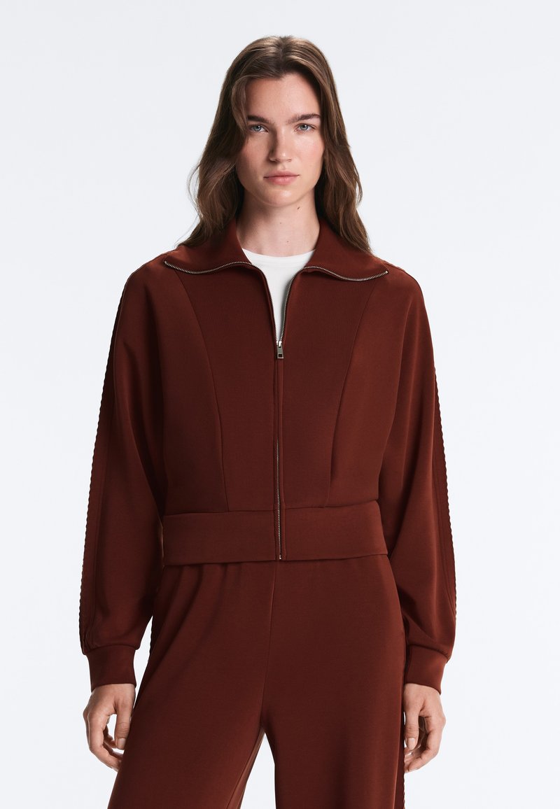 Jeune femme aux longs cheveux bruns porte une veste marron zippée avec manches longues et un pantalon assorti sur un fond clair uni.