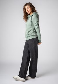 Groene hoodie met een voorzak en een trekkoord capuchon, gecombineerd met zwarte wijde jeans en witte sneakers. Tekstprint op de mouwen en borst.