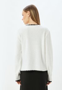 Witte gebreide trui met een gestructureerde afwerking, geaccentueerd door zwarte franjen aan de zoom en mouwcoupons, met een ronde halsontwerp.
