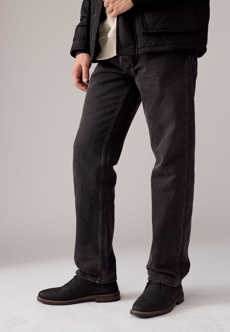 Schwarze Denim-Jeans mit geradem Schnitt und subtiler Textur. Kombiniert mit schwarzen Stiefeletten mit brauner Sohle.