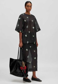Schwarzes besticktes Outfit mit floralen Mustern, transparenter Textur, lockerer Passform, kombiniert mit einer glänzenden schwarzen Tote-Bag und flachen schwarzen Schuhen.