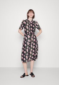 Vestido floral de botão com mangas curtas, fundo preto, padrões de flores rosa e verdes, cintura ajustada e sapatos pretos planos-slip-on.