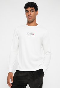 Bijela majica dugih rukava sa crveno-plavim otisnutim logotipom centriranim na prsima. Materijal izgleda mekano; izrez na vratu je okrugao, a rukavi su uski.