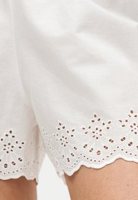 Short en coton blanc avec broderie œillets florale le long de l'ourlet festonné, porté sur la partie supérieure de la cuisse d'une personne.