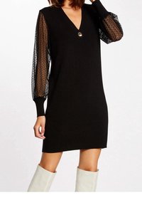 Robe noire avec un décolleté en V, dotée de manches bouffantes en dentelle noire transparente. Agrémentée d'un bouton rond au niveau du col. Tissu en maille douce.