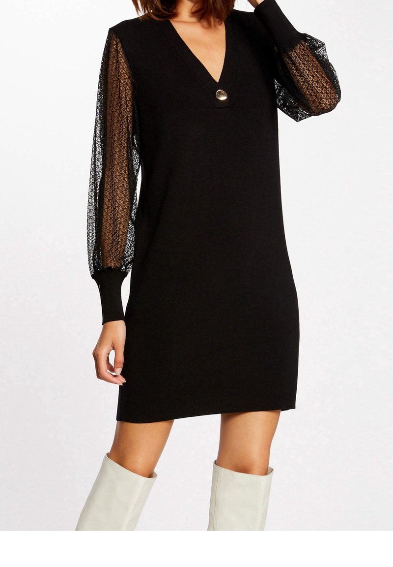 Robe noire avec un décolleté en V, dotée de manches bouffantes en dentelle noire transparente. Agrémentée d'un bouton rond au niveau du col. Tissu en maille douce.