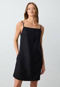 Robe en lin noir avec un décolleté carré et de fines bretelles spaghetti. Elle possède des poches sur les côtés et une silhouette droite jusqu'aux genoux.