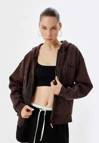 Felpa marrone con zip, realizzata in cotone spesso, con una stampa grafica sul lato sinistro, abbinata a un top corto nero e a pantaloni corti neri.