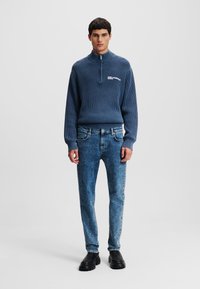 Pull texturé bleu marine avec col montant et fermeture éclair, associé à un jean délavé bleu clair et des chaussures noires. Présente un logo sur le devant du pull.