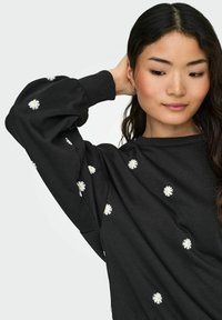 Jauna sieviete ar garām tumšām matēm, tērpta melnā džemperī ar baltām margrietiņu apdrukām, pacēlusi vienu roku aiz galvas.