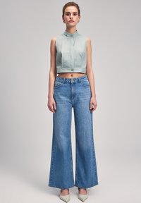Top sin mangas a rayas verde menta, con cuello y cortado, combinado con pantalones vaqueros de mezclilla azul claro de pierna ancha. Detalle de botón en forma de ojal en la parte delantera.