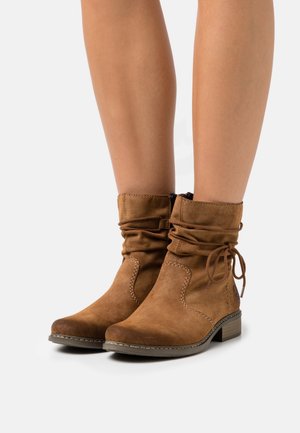 Bottes de cheville en daim marron avec fermeture éclair sur le côté, détail de lien, petit talon empilé, portées sur des jambes nues sur fond blanc.