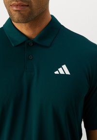 Tyrkysové polo tričko z hladkej tkaniny s preloženým golierom, dvoma gombíkmi a bielym logom s tromi pruhmi na ľavej hrudi.