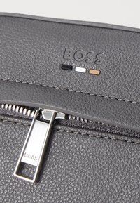 Borsa in pelle grigia con texture, dotata di una cerniera argentata con logo e tre linee di accento colorate: nera, bianca e marrone.