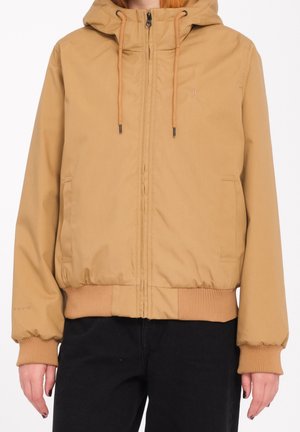 Persona che indossa una giacca con cappuccio color sabbia con polsini e orlo a coste, cerniera frontale, coulisse, tasche laterali e pantaloni neri.