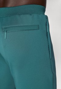Teal treningsbukser med glatt tekstur, elastisk midjebånd og en glidelåslomme på baksiden for sikker oppbevaring.
