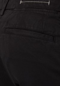 Černé kalhoty s zadní kapsou, jemnou texturou a kontrastním prošíváním nad kapsou. Jednoduchý, funkční design.