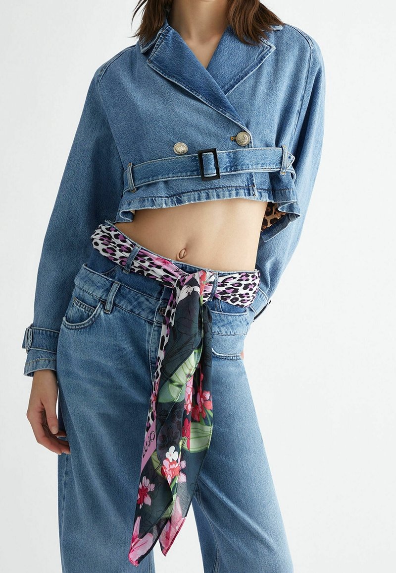 Femme portant une veste en jean courte bleue et un jean taille haute avec une ceinture foulard à motifs floraux et léopard nouée à la taille.