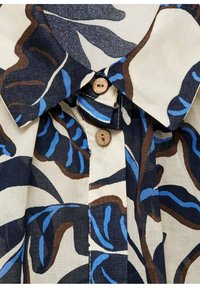 Chemise à col avec motifs abstraits de feuilles bleues, marron et noires sur tissu crème, dotée de deux boutons en bois attachés au col.