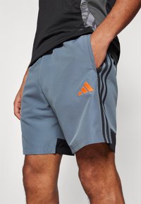 Grå treningsshorts med en oransje Adidas-logo, med svarte sidestipser og et glatt, lett materiale.