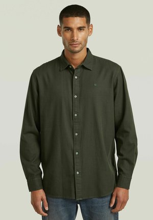 Uomo con capelli corti che indossa una camicia verde oliva a maniche lunghe con bottoni e jeans blu, rivolto verso l'obiettivo su uno sfondo chiaro.