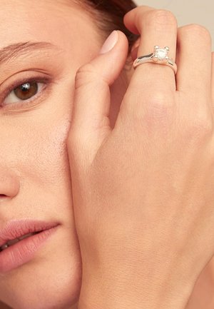 Bague en argent avec une pierre centrale et des accents plus petits, tenue sur une main près du visage, mettant en valeur une peau lisse et un vernis à ongles naturel.