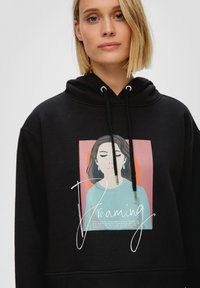 Zwarte hoodie met een grafische print van een vrouw met gesloten ogen en het woord "Dromen" in cursief. De trekkoorden hebben metalen punten.