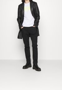 Černý kožený dlouhý kabát přes bílý tričko s neonově žlutým nápisem, doplněný černými slim-fit kalhotami a černými kotníkovými botami s objemnou podrážkou.