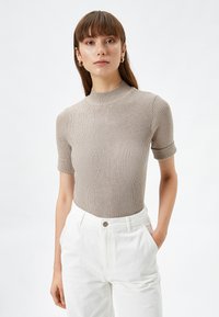 Top de manga corta de canalé beige con cuello alto, combinado con pantalones blancos. La tela tiene una apariencia texturizada y un diseño ajustado.