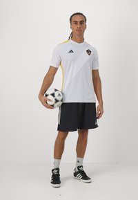 Maglia bianca Adidas con strisce diagonali, pantaloni corti neri e scarpe da ginnastica nere. Il giocatore tiene in mano un pallone da calcio bianco e nero.