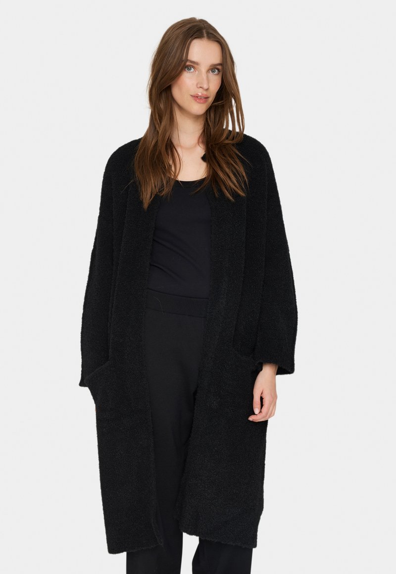 Svart, lång, plysch cardigan med öppen framsida, två framfickor och en mysig textur. Ärmarna är vida och beskurna vid handleden.