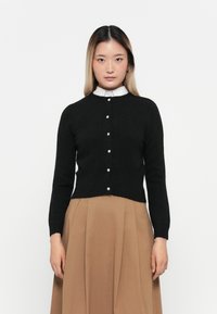 Young woman with straight hair wearing a black buttoned cardigan over a white collared shirt and a high-waisted brown skirt against a white background.