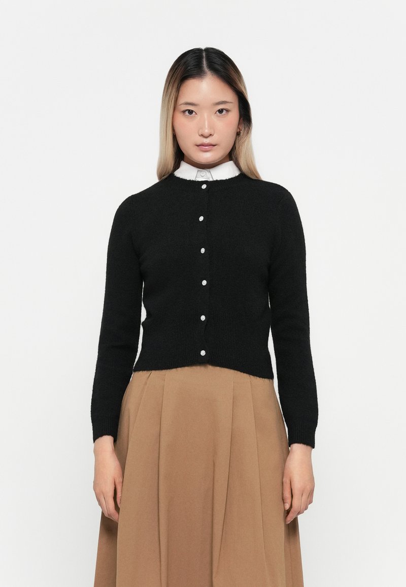Jeune femme aux cheveux raides portant un cardigan noir à boutons sur une chemise blanche à col et une jupe marron taille haute sur fond blanc.