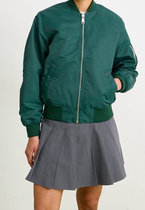 Person trägt eine dunkelgrüne Bomberjacke mit Reißverschluss und einen grauen plissierten Minirock, mit einer Hand in der Jackentasche.