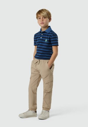 Junge steht mit den Händen in den Taschen, trägt ein blau gestreiftes Poloshirt, beige Cargohosen und weiße Turnschuhe vor einem einfarbigen Hintergrund.