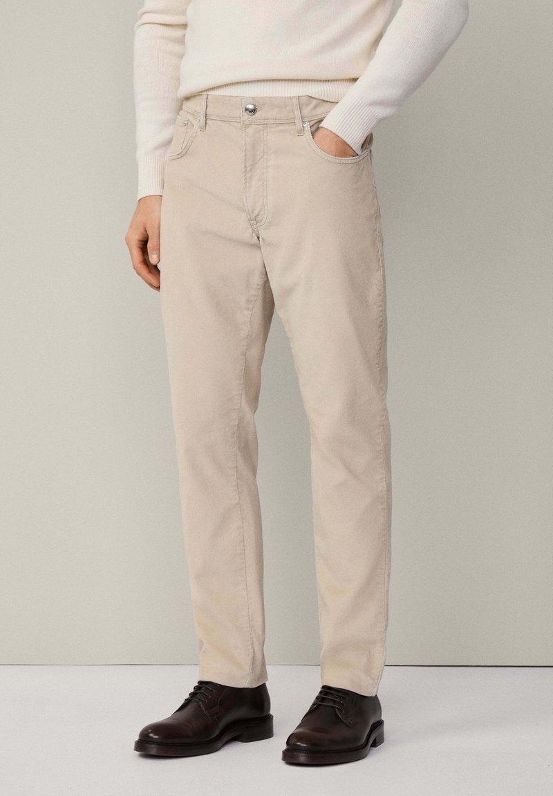 Homme portant un pantalon beige, un pull crème et des chaussures habillées en cuir marron foncé, une main dans la poche, debout devant un fond uni.