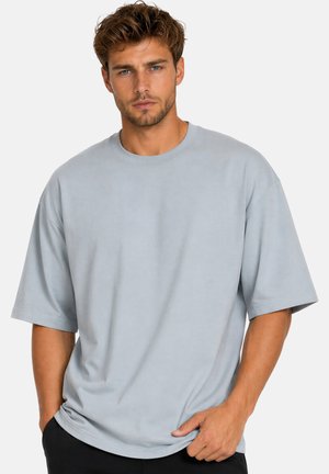 Man die voor een effen achtergrond staat en een loszittend lichtgrijs T-shirt met korte mouwen en een donkere broek draagt, kijkt recht vooruit.