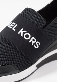 Schwarzer Slip-On-Schuh mit einem strukturierten Mesh-Obermaterial, einem breiten elastischen Band mit der Aufschrift "MICHAEL KORS" in Weiß und einer glänzenden schwarzen Zehenkappe.