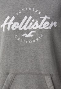 Gray sweatshirt with white embroidered text, featuring a circular logo that includes "Hollister" and "Southern California," and a front pocket.