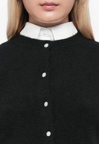 Blonde person wearing black knit cardigan with decorative pearl-like buttons over white collared shirt.