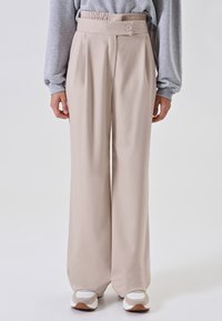 Persona che indossa pantaloni a vita alta beige a gamba larga con una cintura bottonata, abbinati a sneakers bianche e beige, in piedi davanti a uno sfondo bianco.