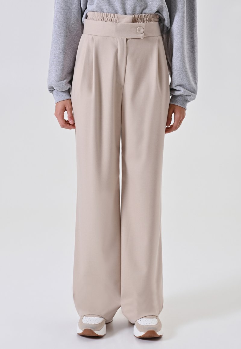 Persona che indossa pantaloni a vita alta beige a gamba larga con una cintura bottonata, abbinati a sneakers bianche e beige, in piedi davanti a uno sfondo bianco.