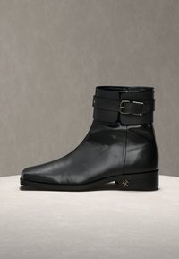 Μαύρο δερμάτινο μποτάκι αστραγάλου με μυτερή μύτη, χαμηλό τακούνι, φαρδιά λουρίδα και μεταλλική αγκράφα στην κορυφή, παρουσιασμένο σε ουδέτερη επιφάνεια.